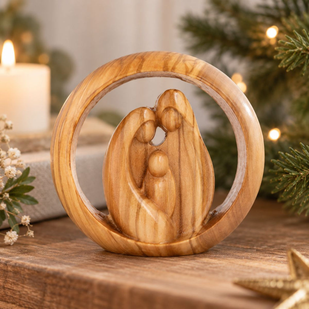 A hand-carved olive wood figurine of Mary, Joseph, and baby Jesus, set inside a circular olive wood frame. The sculpture is displayed on a wooden surface with a softly blurred Christmas background featuring a lit candle, pine branches, and warm string lights.
