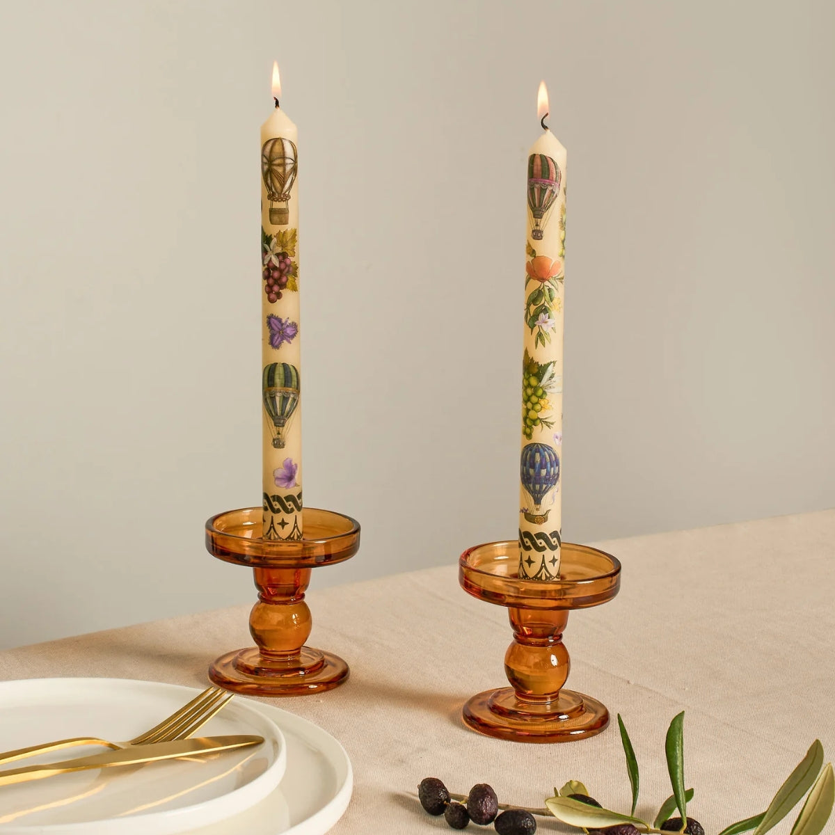 Two lit cream taper candles with colorful hot air balloon and fruit illustrations, displayed in amber glass candle holders on a beige tablecloth with plates and olive branches nearby.