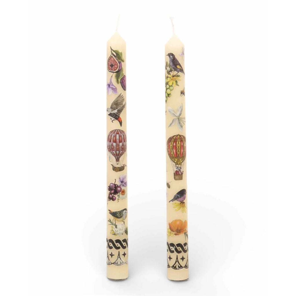 Two cream-colored taper candles with vintage-style illustrations of birds, hot air balloons, figs, and grapes on a white background.