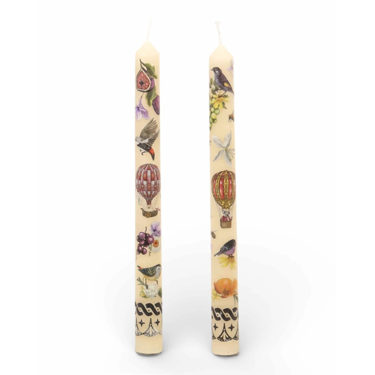 Two cream-colored taper candles with vintage-style illustrations of birds, hot air balloons, figs, and grapes on a white background.