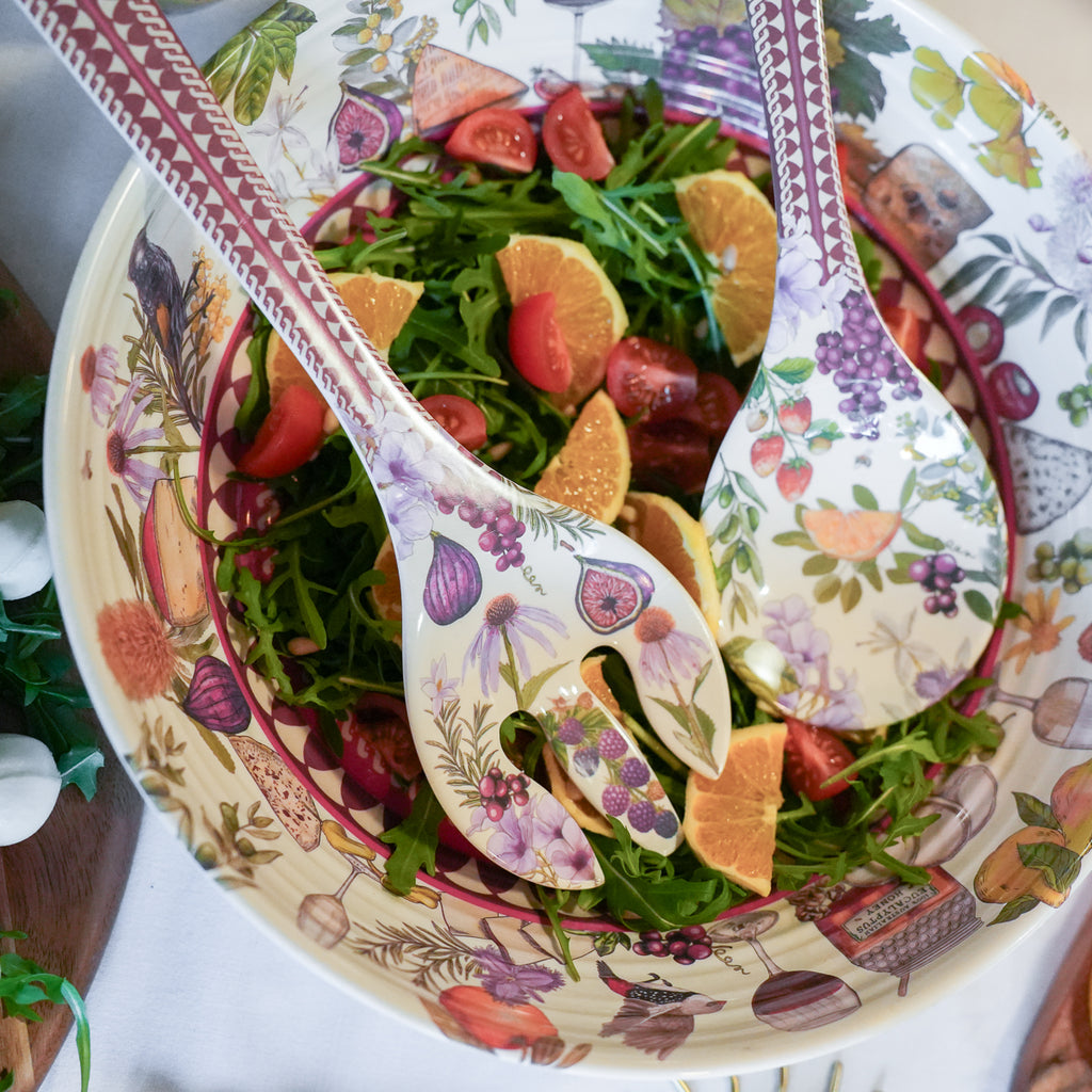 A top-down view of a decorative salad bowl filled with greens, arugula, figs, tomatoes, and orange slices, with two matching patterned salad spoons resting in the bowl.