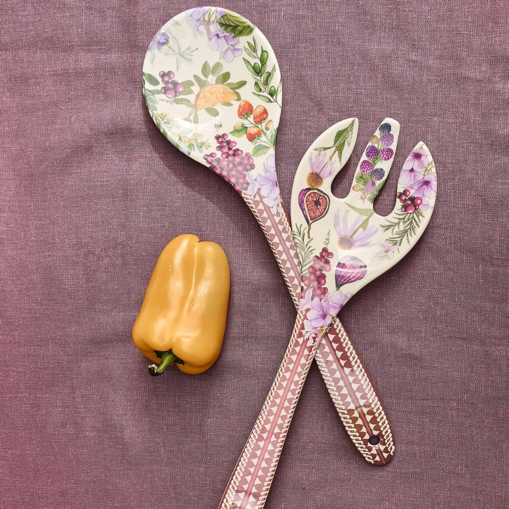 Two decorative serving utensils, a spoon and a salad fork, with a floral fruit print and pink patterned handles, arranged on a textured mauve fabric next to a yellow bell pepper.