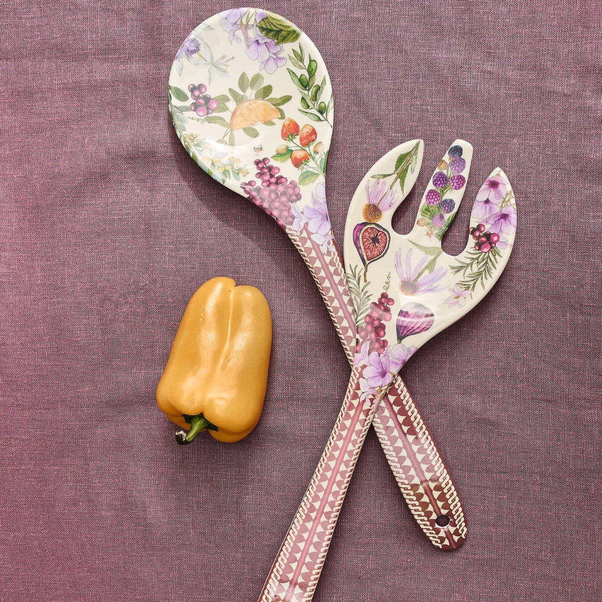 Two decorative serving utensils, a spoon and a salad fork, with a floral fruit print and pink patterned handles, arranged on a textured mauve fabric next to a yellow bell pepper.