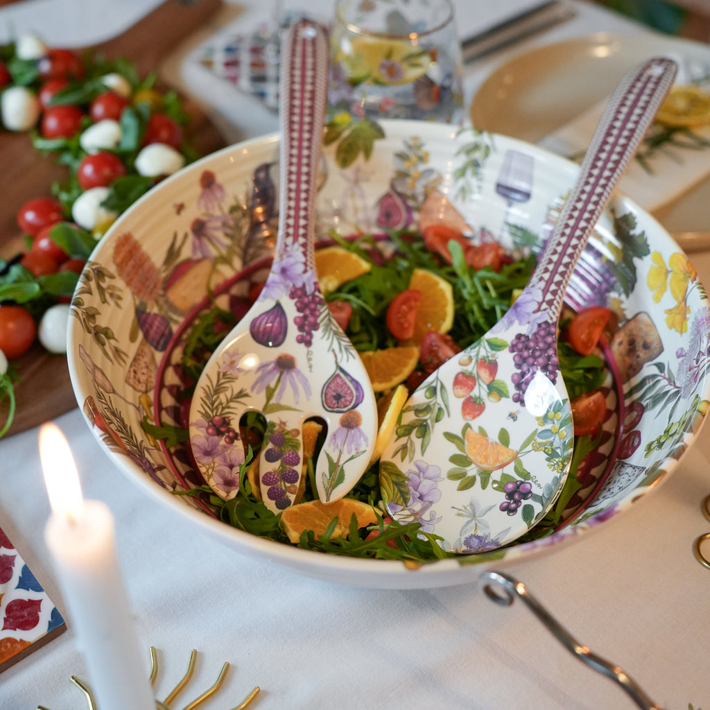 A large ceramic salad bowl filled with greens, figs, tomatoes, and citrus, with two matching patterned salad spoons resting inside. The bowl and spoons feature a colorful botanical print with purple grapes, orange slices, and floral motifs on a white base.