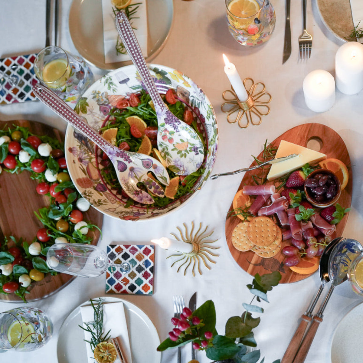 An overhead view of a beautifully set dining table for a holiday feast, featuring a roasted turkey on a platter, a large salad bowl, a charcuterie board, a bottle of red wine, candles, and place settings on a white tablecloth.