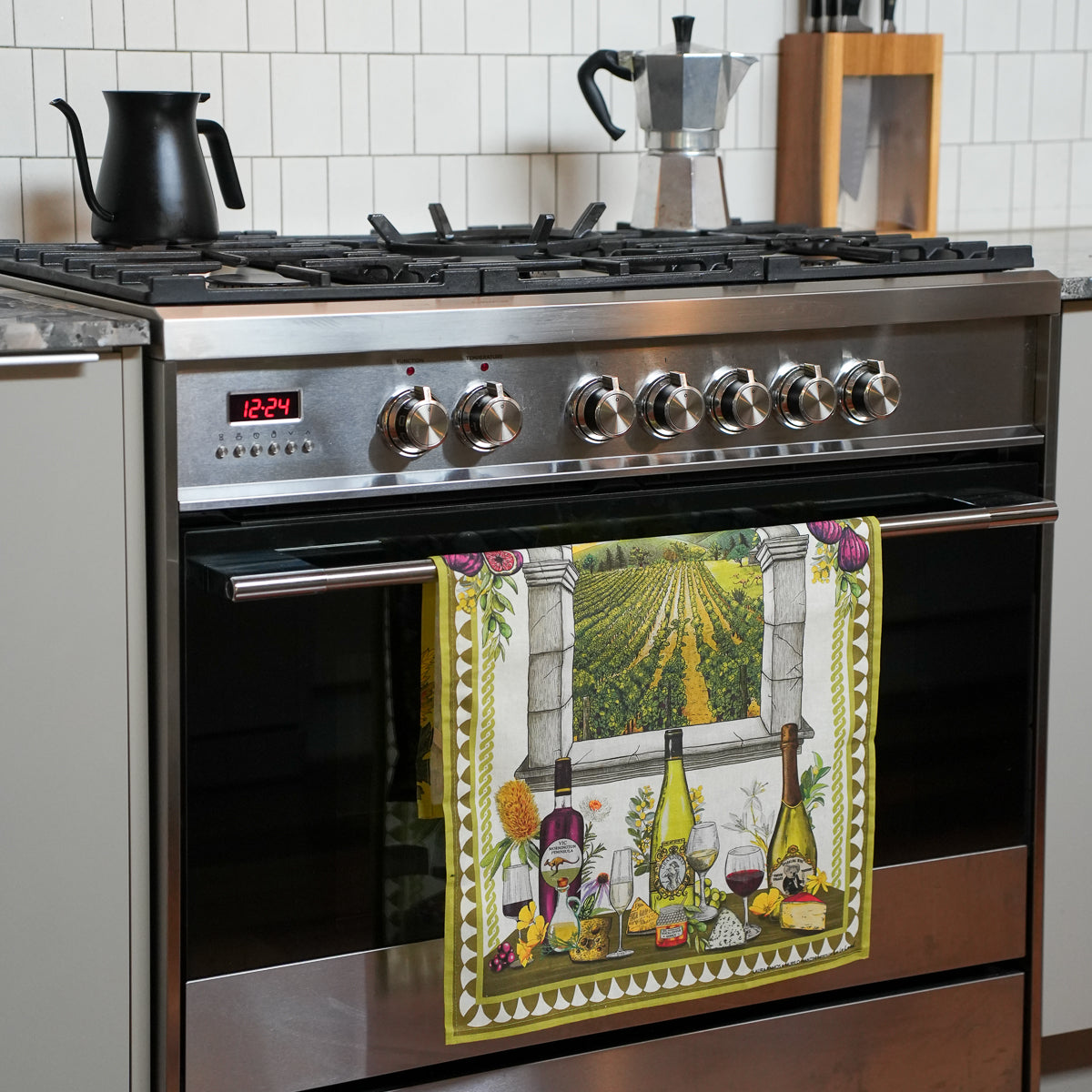 A kitchen towel with a wine-themed vineyard illustration hangs on the handle of a stainless steel gas range in a kitchen setting.