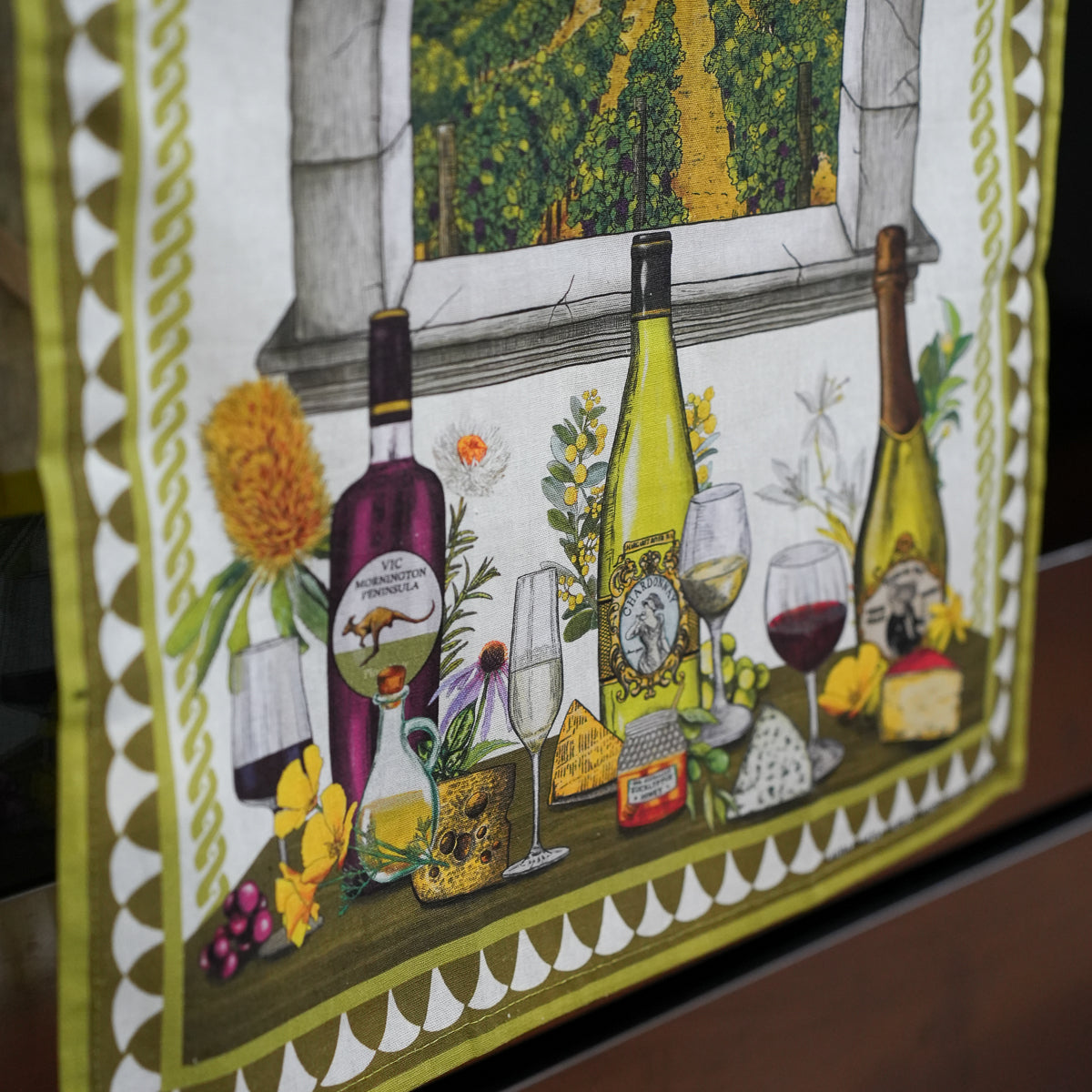 A close-up of a decorative textile or wall hanging featuring an illustrated wine-themed scene with bottles, wine glasses, cheese, flowers, and a vineyard window view. The fabric has a yellow-green border and is photographed against a dark real-world background.