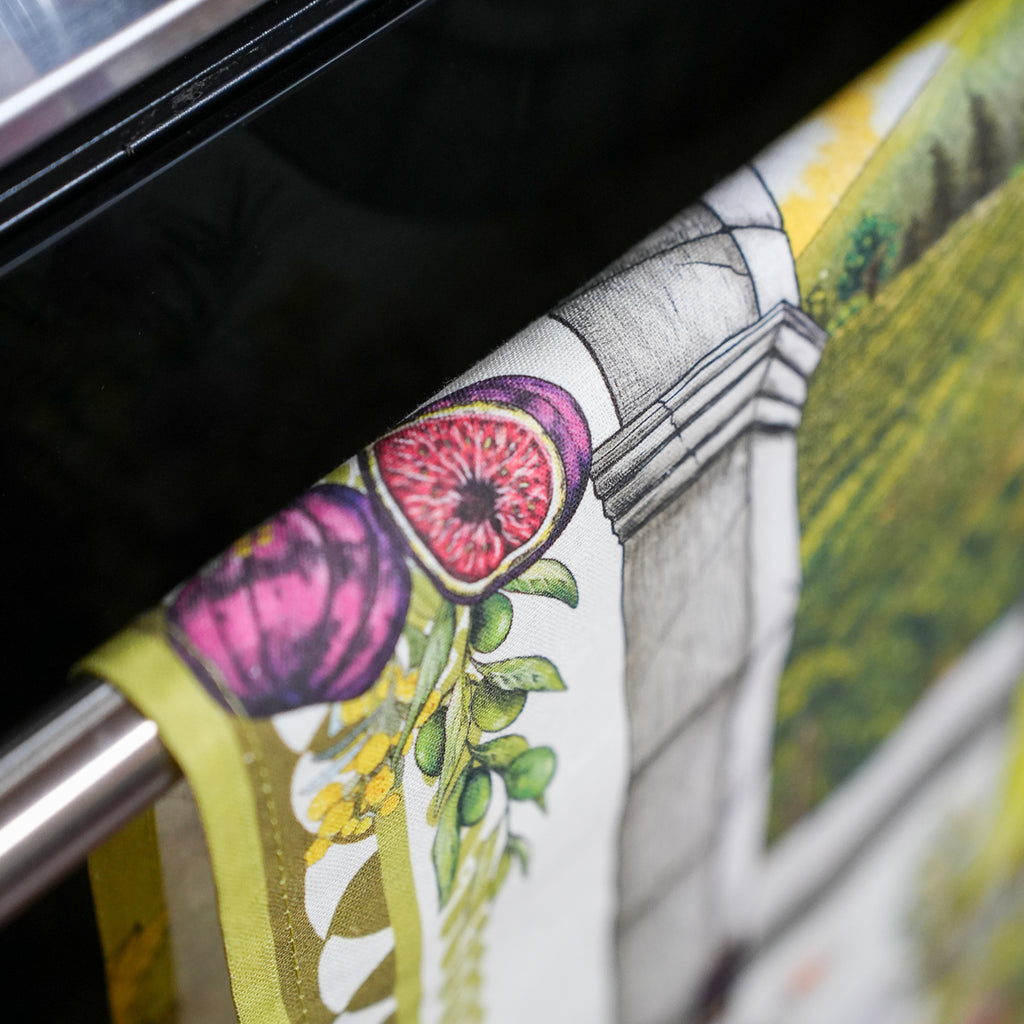 A close-up of a printed textile draped over a metal rod, featuring a botanical and architectural design with pink figs, green leaves, and a gray column on a white background.