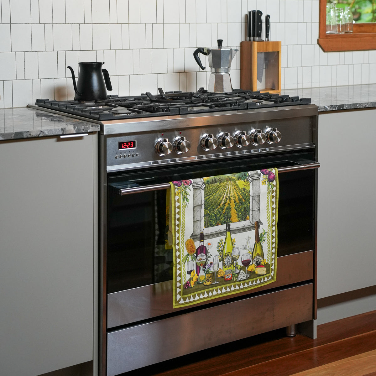 A stainless steel gas range oven in a modern kitchen, with a colorful decorative kitchen towel hanging from the handle. The towel features a yellow border and an illustration of a table with wine bottles, glasses, and a basket under a window.