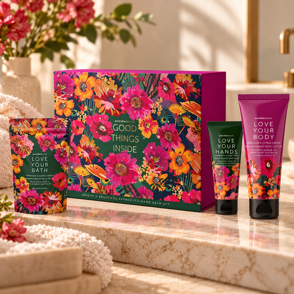 A bath and body gift set displayed on a marble surface with flowers and towels in a bathroom setting. The set includes a large floral gift box labeled 'Good Things Inside,' a pouch labeled 'Love Your Bath,' a tube labeled 'Love Your Hands,' and a squeeze bottle labeled 'Love Your Body.