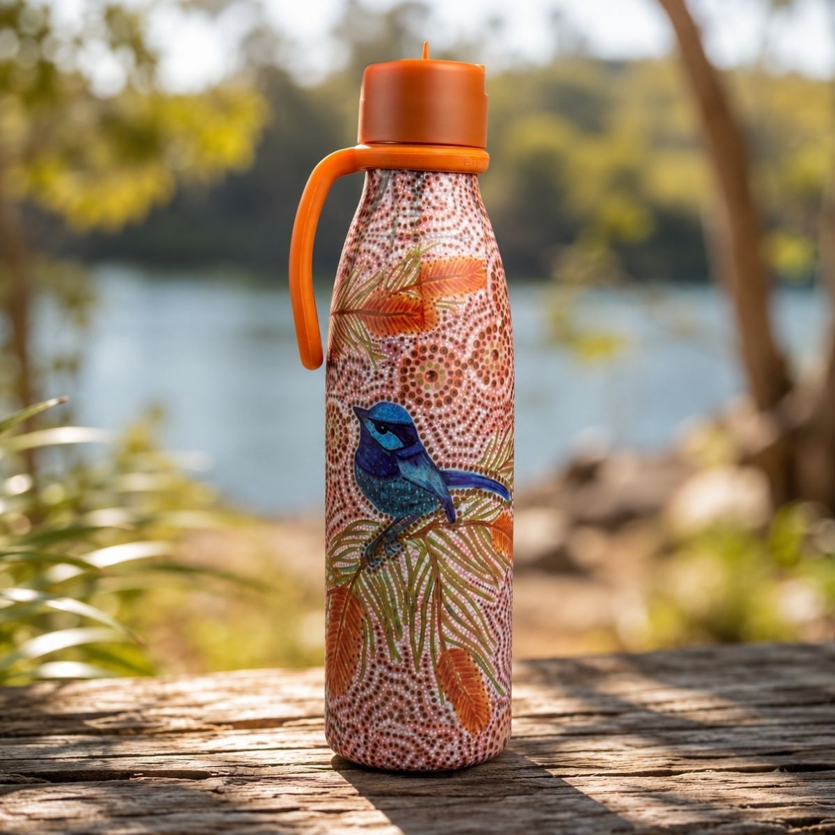 A reusable insulated bottle with a bright orange lid and handle, decorated with a red, orange, and blue dot-art style pattern featuring a blue bird, standing on a wooden surface outdoors with a blurred lake and trees in the background.