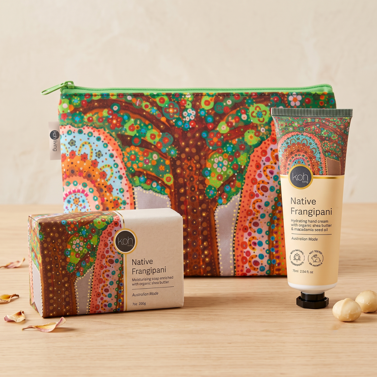 A set of Native Frangipani self-care items from the brand KooKoo, including a hand cream tube, a soap bar, and a zippered pouch, all decorated with a vibrant Aboriginal dot art pattern, arranged on a light wooden surface.