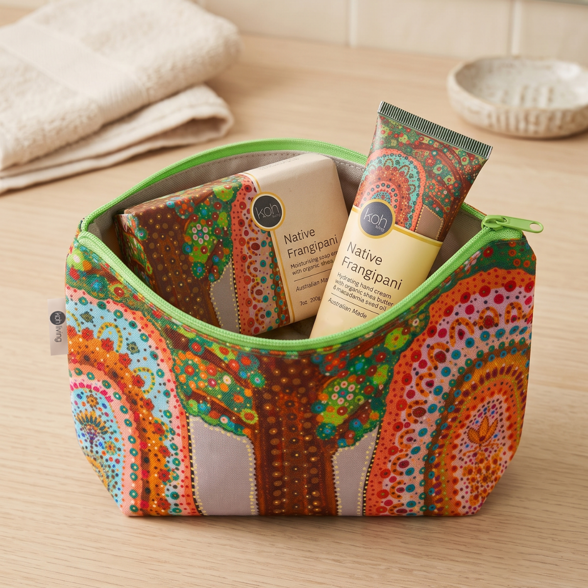 A patterned cosmetic pouch with a green zipper sits on a wooden surface, filled with Native Frangipani bath products. The pouch features a vibrant, multicolored mandala-style tree design in orange, green, brown, and blue.