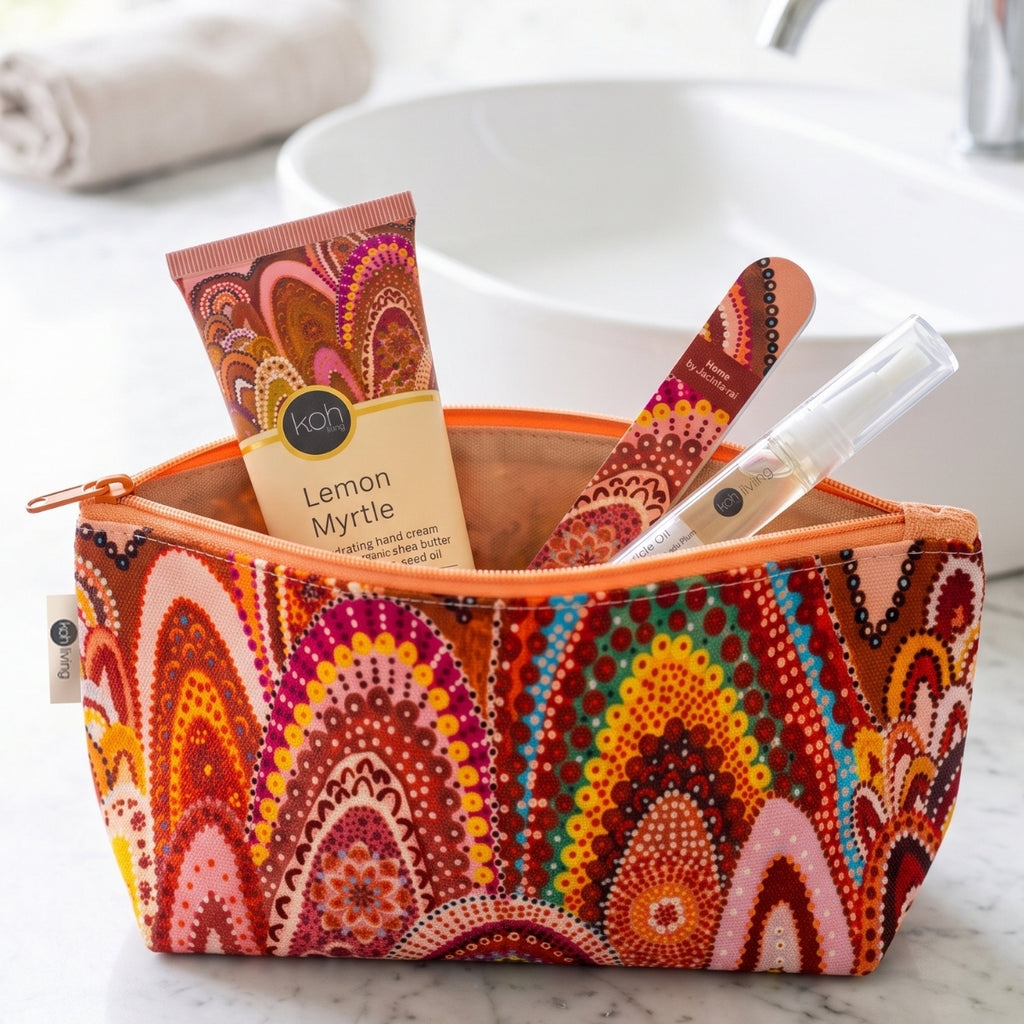 A colorful patterned toiletry bag sits on a marble countertop with a sink and towel blurred in the background. Inside the bag are a tube of Kon Lemon Myrtle hand cream, a wooden nail file, and a clear lip balm tube.