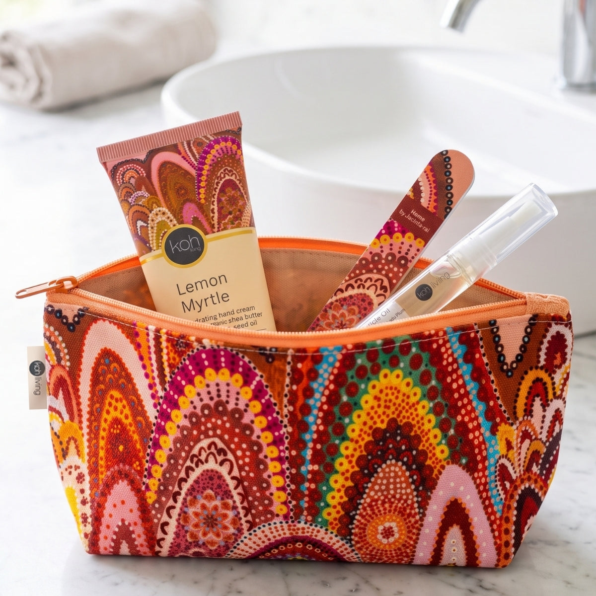 A colorful patterned toiletry bag sits on a marble countertop with a sink and towel blurred in the background. Inside the bag are a tube of Kon Lemon Myrtle hand cream, a wooden nail file, and a clear lip balm tube.