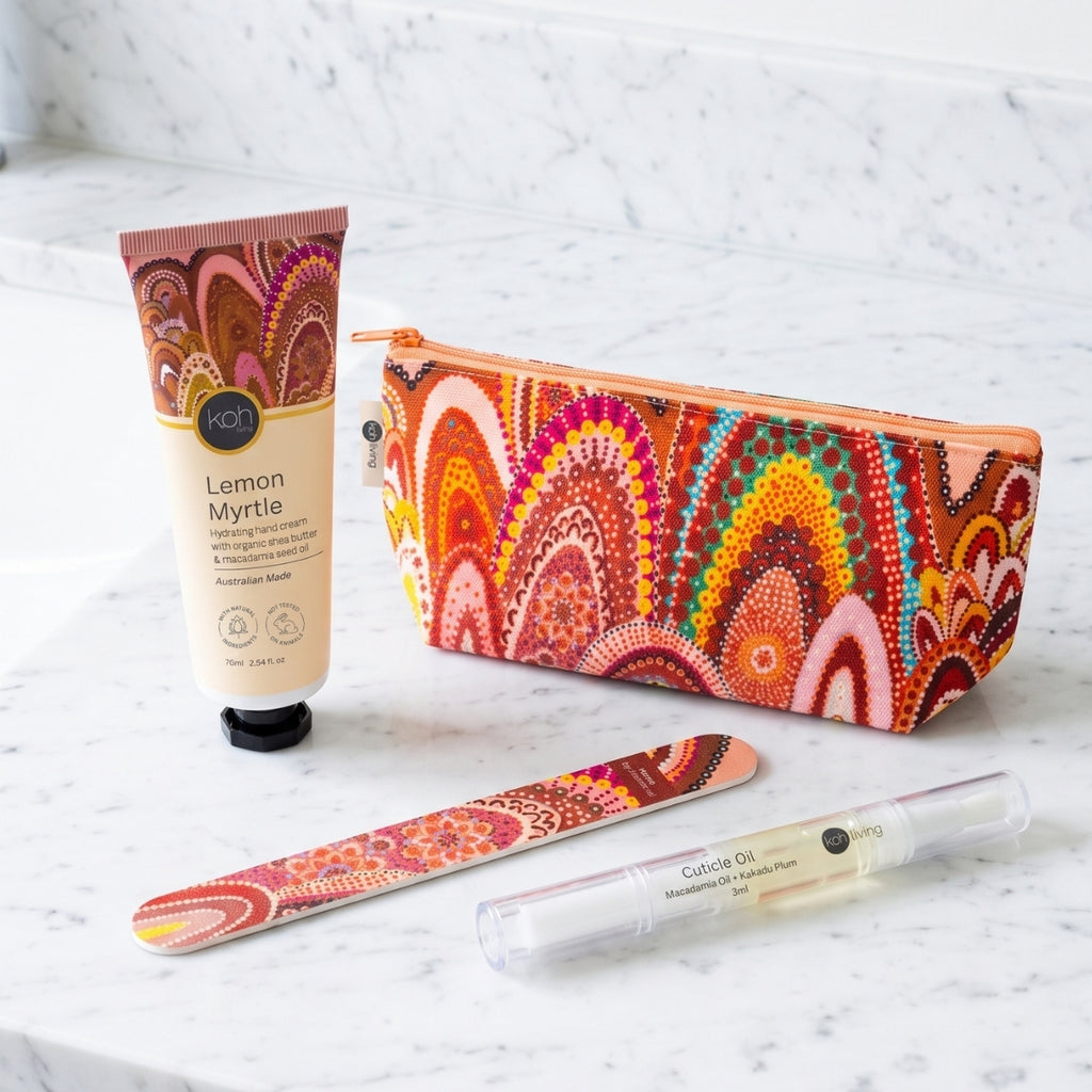 A coordinated set of beauty and personal care items arranged on a white marble surface: a tube of AOT Lemon Myrtle hand cream, a zip pouch, a nail file, and a clear lip balm tube. The pouch, nail file, and tube feature a bold multicolor abstract pattern in orange, red, pink, yellow, green, and brown tones, while the hand cream tube is cream with a brown cap.