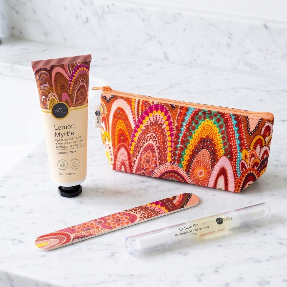 A coordinated set of beauty and personal care items arranged on a white marble surface: a tube of AOT Lemon Myrtle hand cream, a zip pouch, a nail file, and a clear lip balm tube. The pouch, nail file, and tube feature a bold multicolor abstract pattern in orange, red, pink, yellow, green, and brown tones, while the hand cream tube is cream with a brown cap.