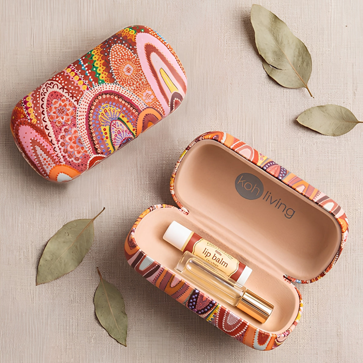 An open and closed matching travel lip balm case set on a light beige textured surface with scattered dried leaves. The cases feature a vibrant, multicolored bohemian paisley pattern in orange, pink, red, yellow, and blue.
