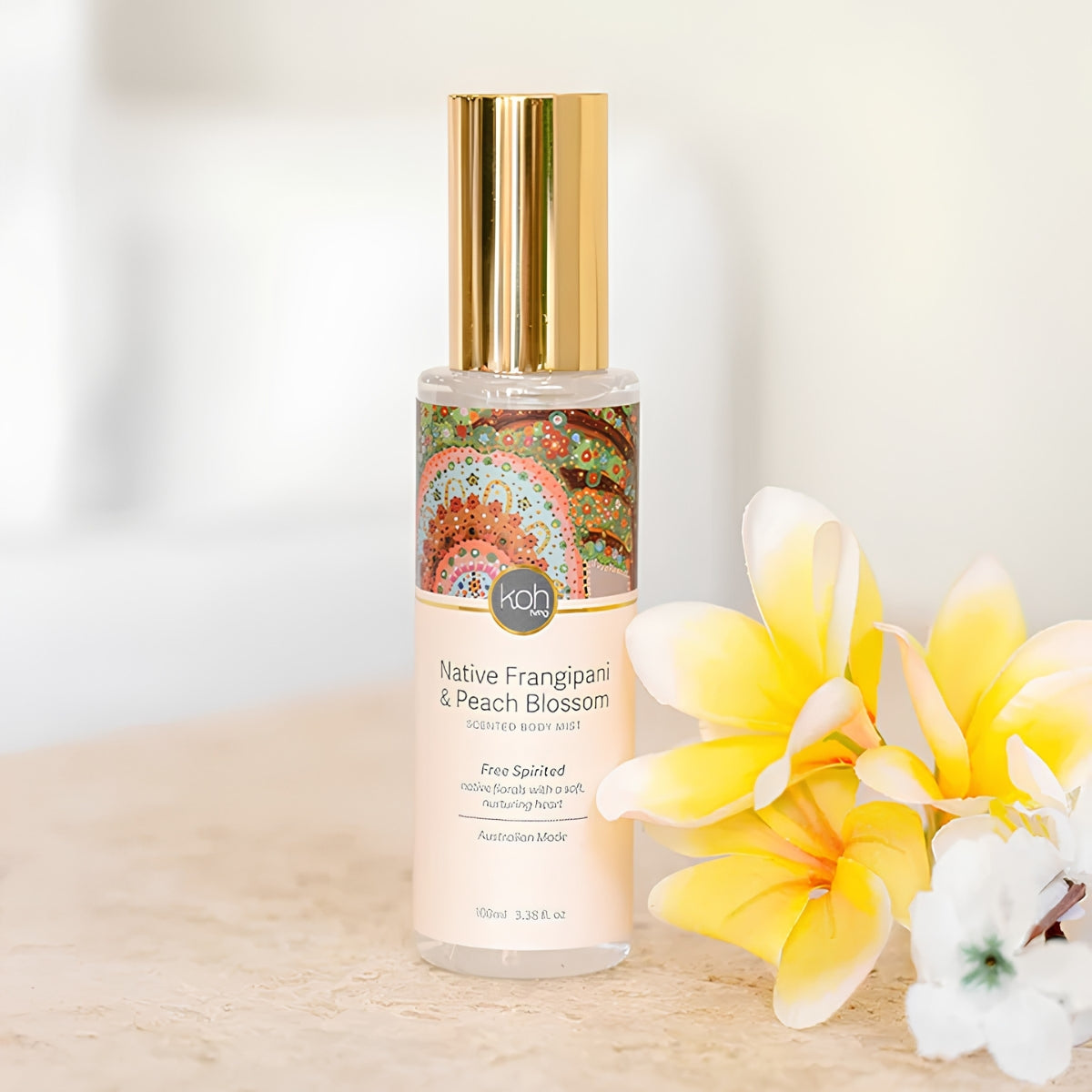 A bottle of Native Frangipani & Peach Blossom Recycled Body Mist by KOH, featuring a gold cap and a label with a colorful mandala design, styled with yellow and white plumeria flowers on a light beige surface.