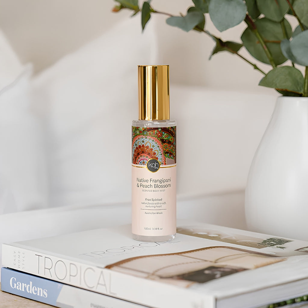 A bottle of body mist Native Fragrance & Peach Blossom pure scented with a gold cap and peach-colored label, styled on a stack of books next to a white vase with green leaves.