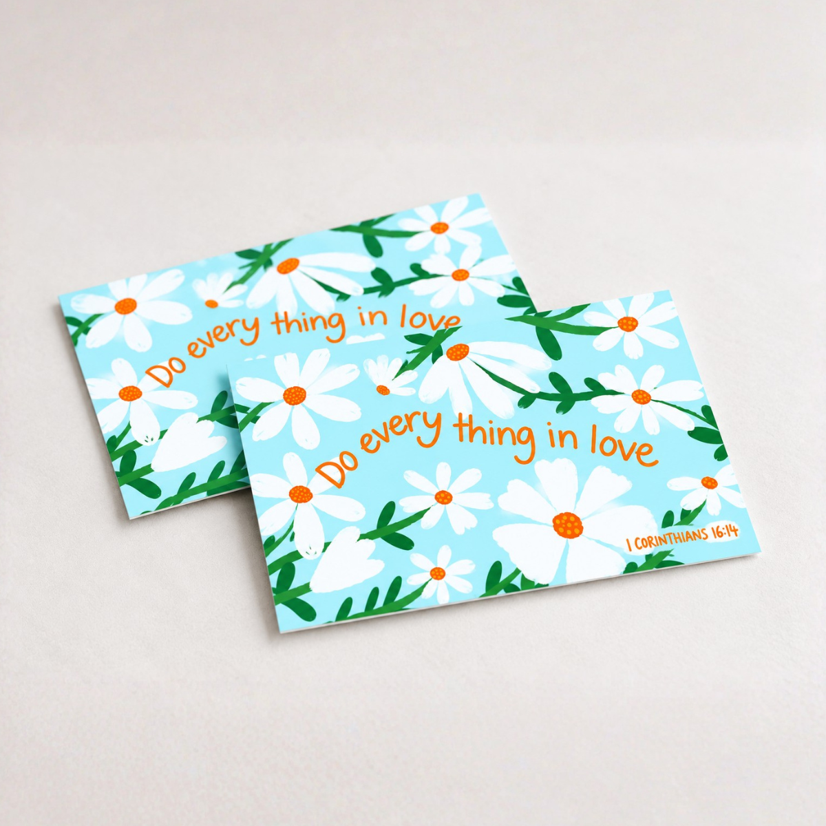 Two square cards with a light blue background, white daisy flowers, green leaves, and orange text reading 'Do every thing in love' and '1 Corinthians 16:14', shown on a light neutral surface.