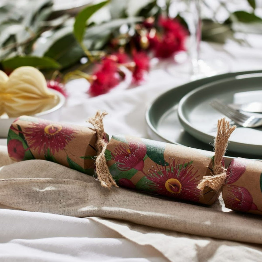 A close-up of a cracker-style party favor wrapped in brown kraft paper with a pink and green floral print, tied with twine on a styled table setting with plates, leaves, and red flowers in the background.
