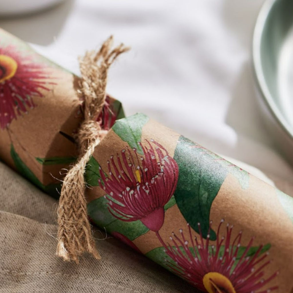 A rolled kraft paper package wrapped with a botanical print of pink flowers and green leaves, tied with a rustic jute twine bow, set on a neutral fabric surface.