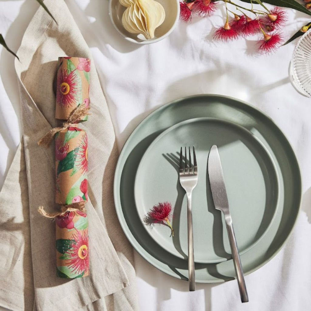 A styled tabletop flat lay showing a floral paper cracker tied with twine, a sage green plate, silver fork and knife, and pink flowers on a white cloth background.