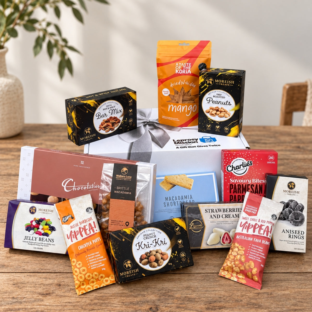 A curated gourmet gift hamper arranged on a wooden table, featuring an assortment of packaged snacks, chocolates, dried fruits, and a white gift box tied with a gray ribbon. Visible brands include Morriston, Appt.