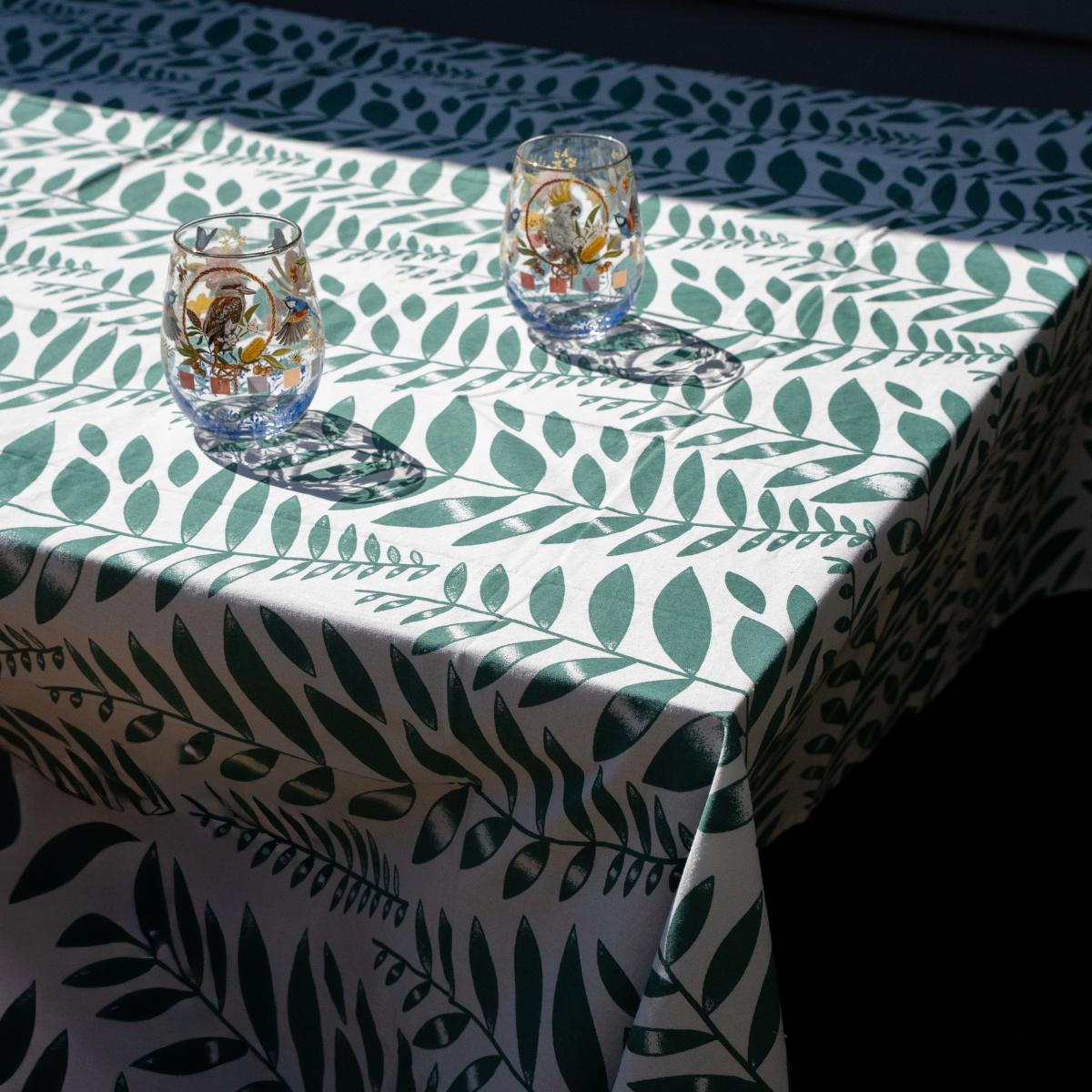 A white tablecloth with a green leaf pattern draped over a table, with two clear glass candle holders placed on top in bright sunlight.