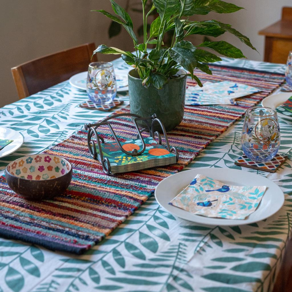 A styled dining table setting featuring a colorful woven striped placemat, a green potted plant, patterned glass goblets, white plates with floral napkins, and a matching leaf-print tablecloth.