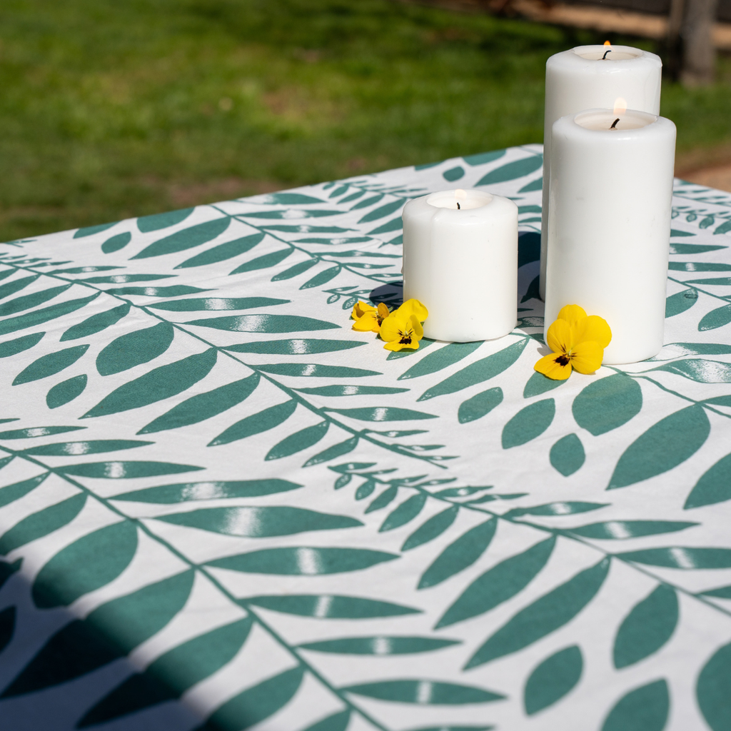 A white tablecloth with a green leaf pattern is draped over an outdoor table, styled with three white pillar candles and small yellow flowers, with a blurred green lawn in the background.