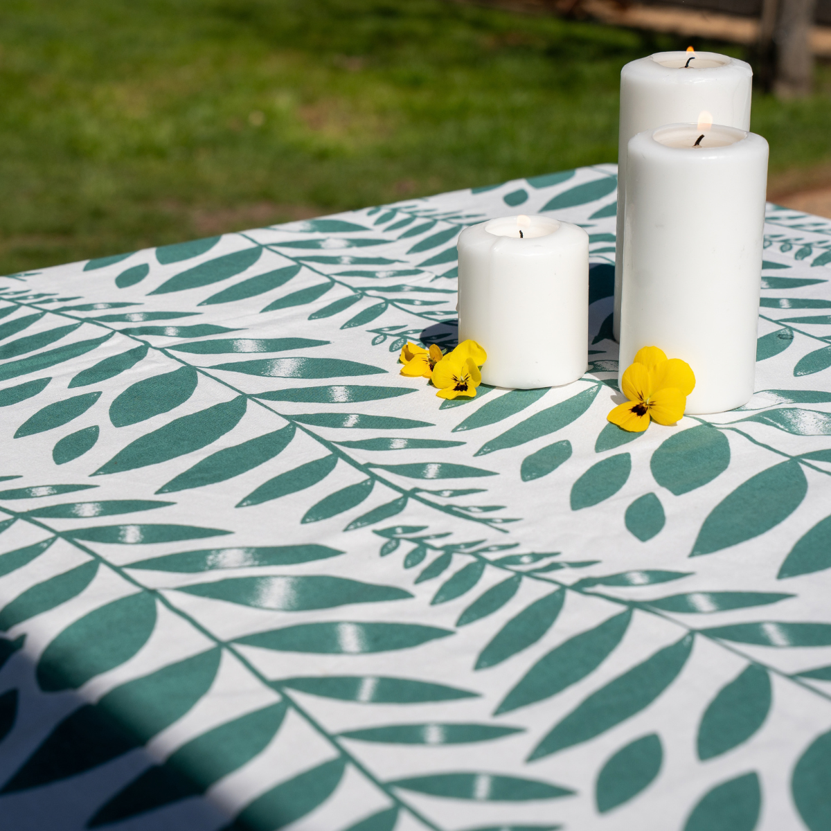 A white tablecloth with a green leaf pattern is draped over an outdoor table, styled with three white pillar candles and small yellow flowers, with a blurred green lawn in the background.