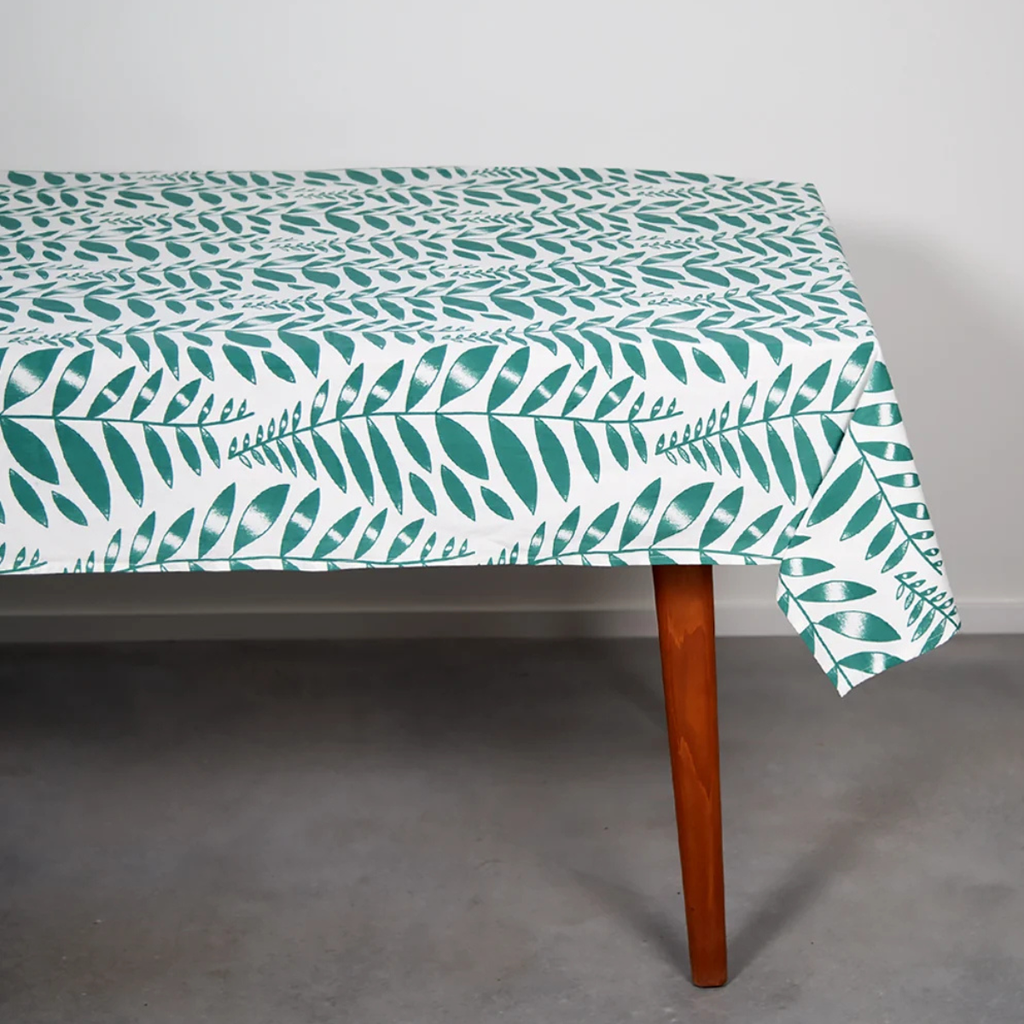 A rectangular table covered with a white tablecloth featuring a repeating teal-green leaf pattern, shown with one wooden leg visible against a plain light gray wall and concrete floor.