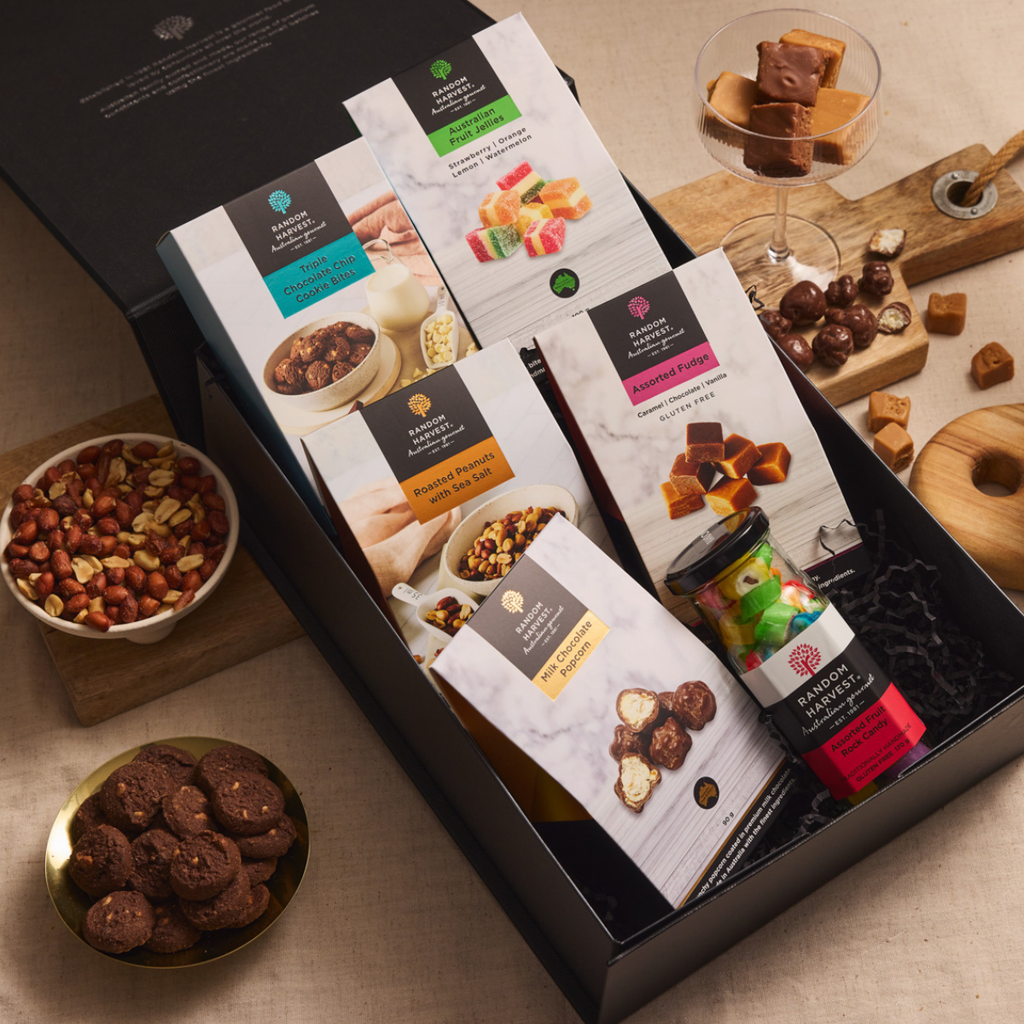 A gourmet gift box of chocolate and candy treats, featuring packaged snacks like pralines, marshmallow treats, and chocolate-covered nuts, arranged in a black box with decorative bowls of ingredients on a beige table.