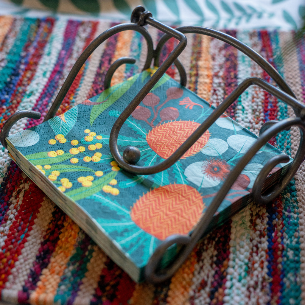 A small notebook with a colorful floral and botanical cover design, resting on a dark metal wire stand over a striped woven textile background.