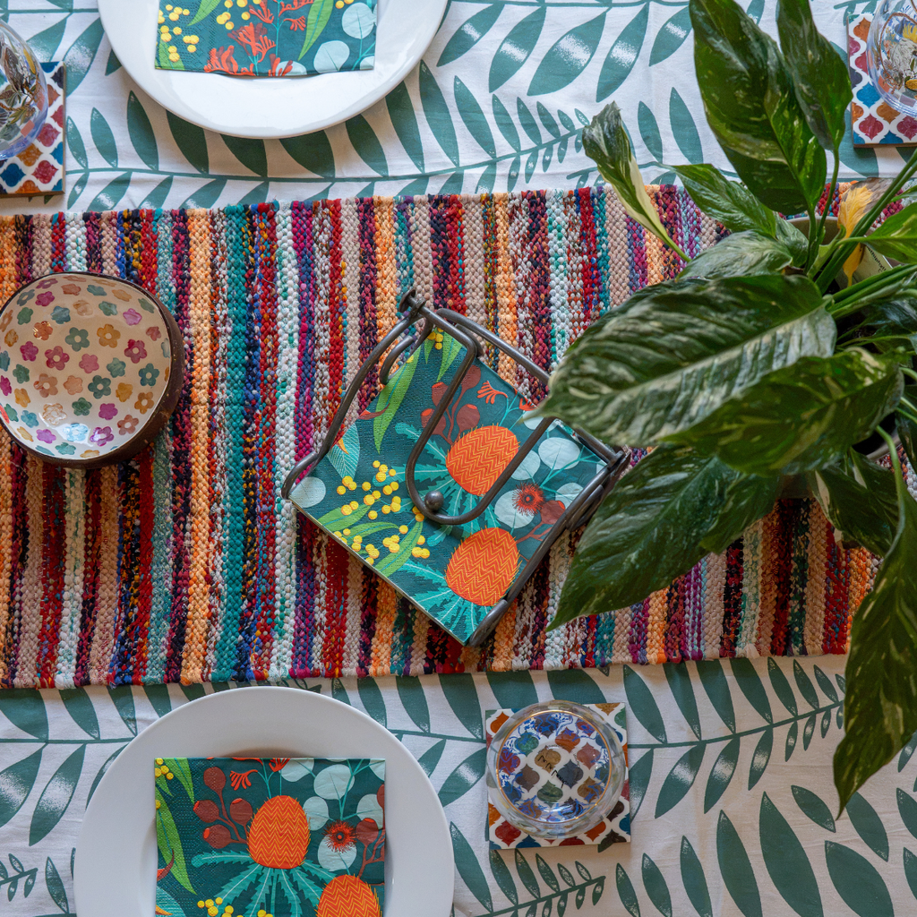 A styled tabletop scene with a colorful woven striped runner, matching patterned napkin, bowls, plates, and a potted green plant on a leaf-patterned tablecloth.