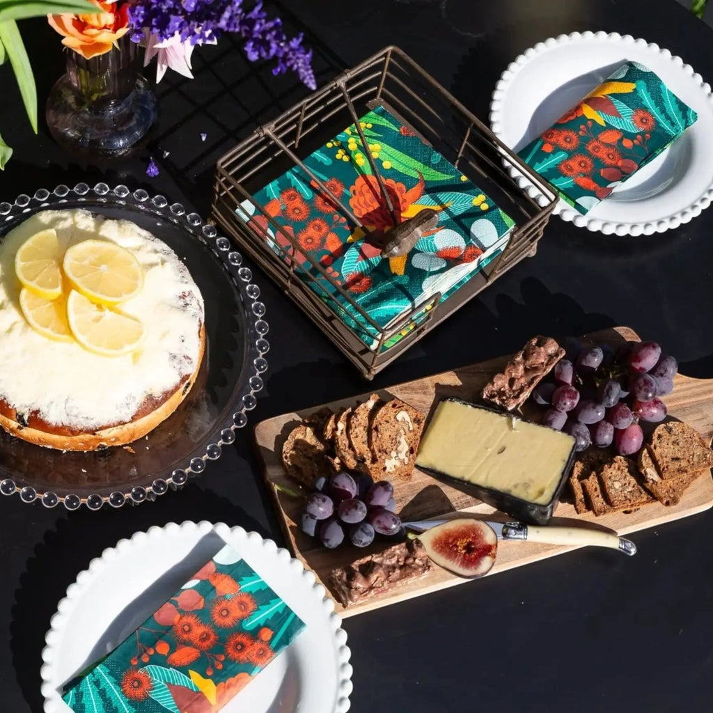 A styled outdoor food table scene with a black table, a decorated cheese cake on a glass plate, a charcuterie board with crackers, grapes, cheese, and a small bottle, plus several colorful patterned napkins and a matching napkin on plates. The product appears to be the napkin set rather than the food.