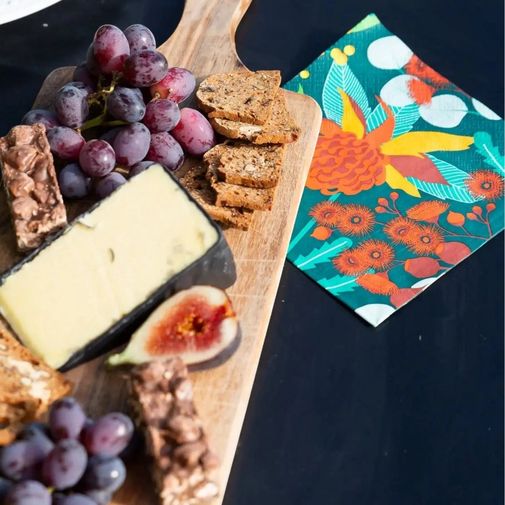 A styled charcuterie board on a dark surface featuring a wedge of cheese, sliced fig, purple grapes, crackers, and nut bars, alongside a colorful floral napkin.