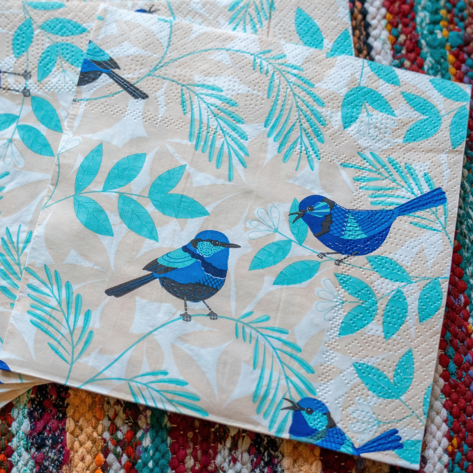 A stack of square paper napkins with a printed pattern of blue birds and teal leaves on a cream background, displayed on a colorful woven rug.