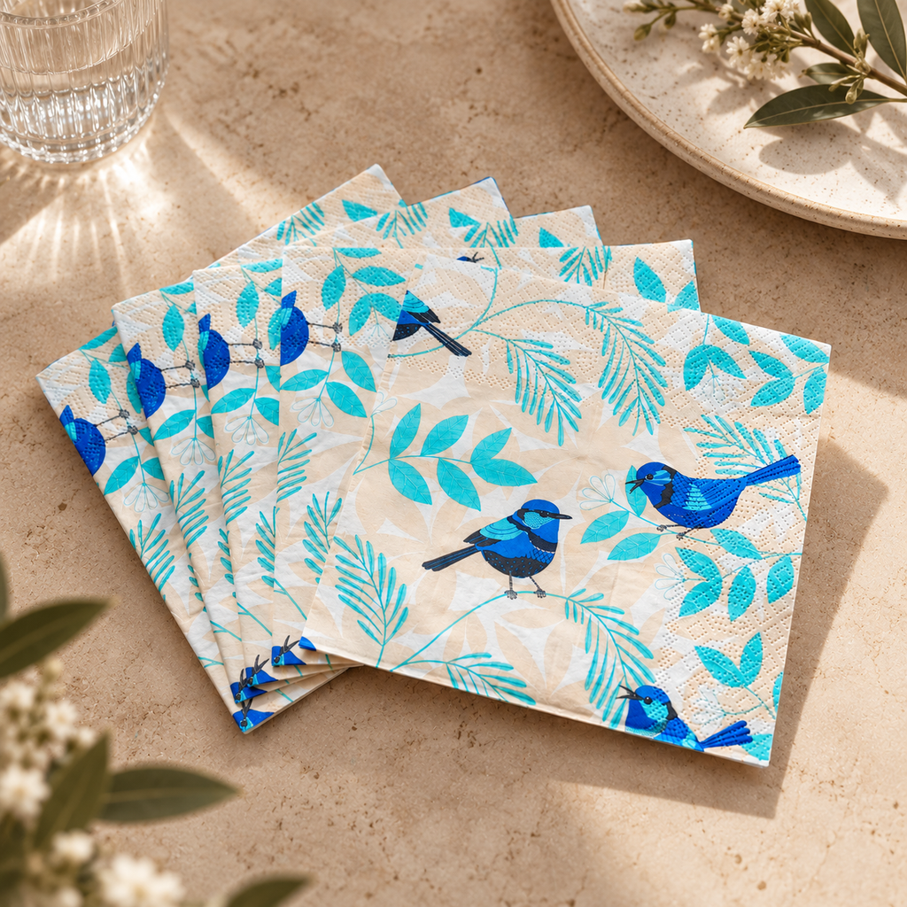 A stack of square paper napkins with a pattern of blue birds and turquoise leaves on a light beige background, styled on a beige tabletop with a glass, plate, and greenery.