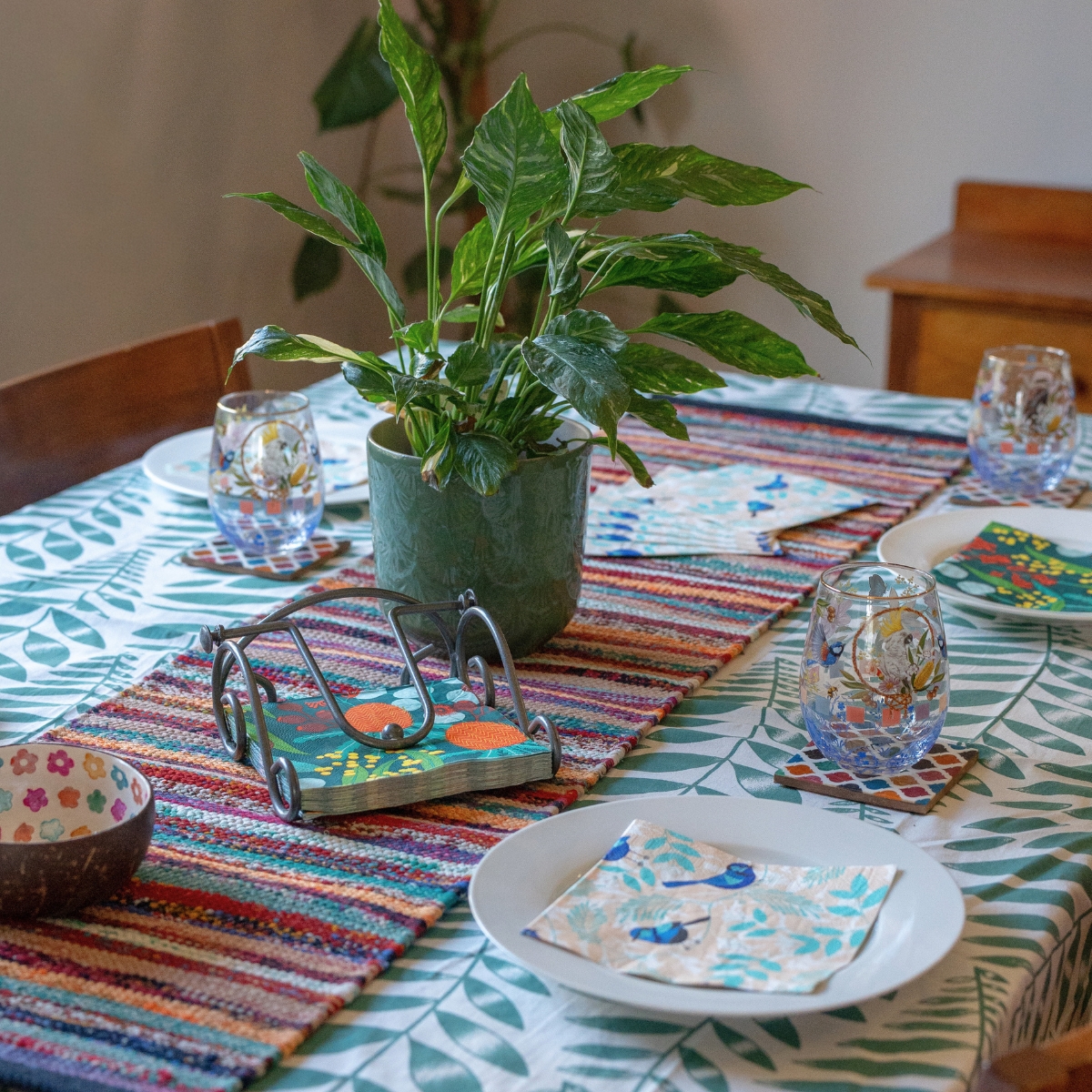 A styled dining table setting featuring a colorful striped woven runner, a green potted plant, patterned napkins, and blue patterned drinking glasses on coasters.