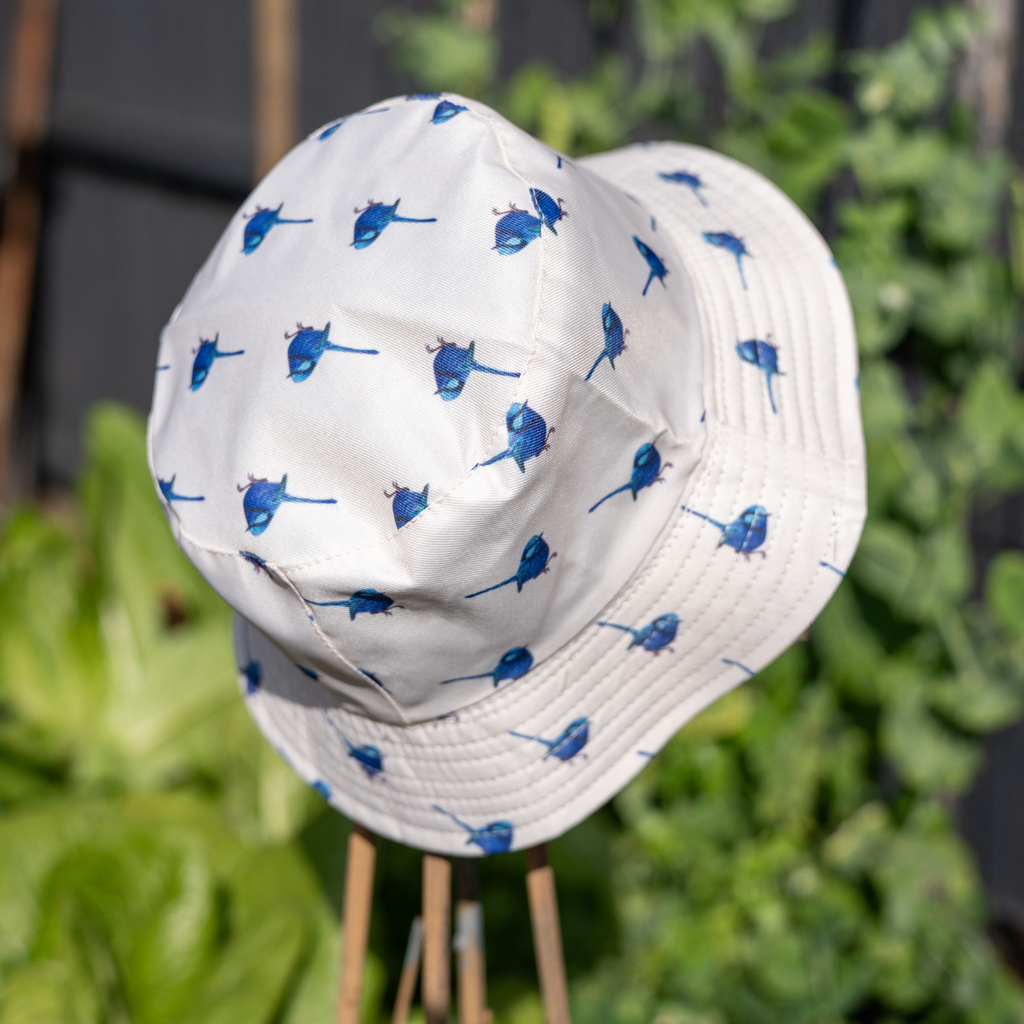 A white bucket hat with a blue bird pattern, displayed on a wooden stand against a blurred green garden background.