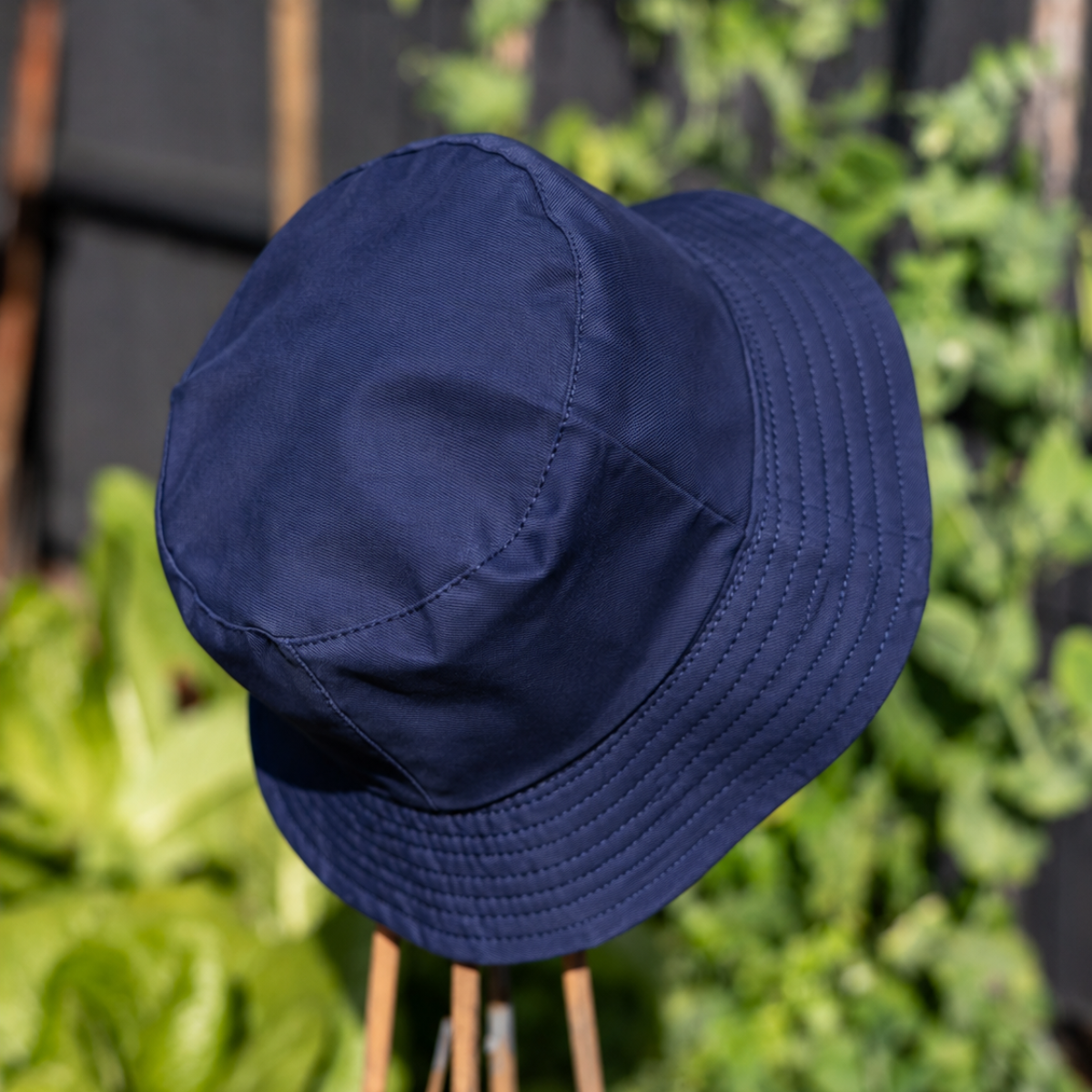A navy blue bucket hat photographed outdoors on a stand, with blurred green foliage and a dark fence in the background.