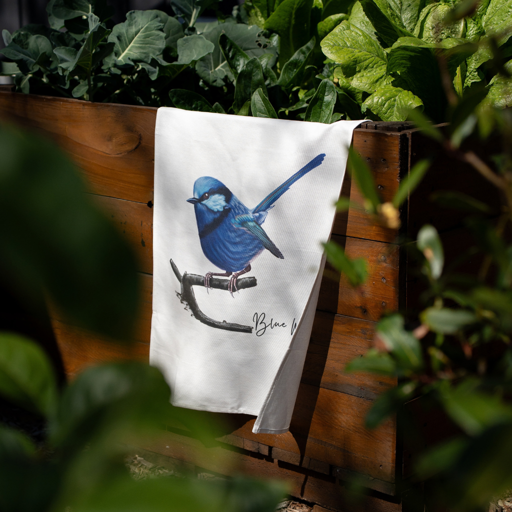A white kitchen tea towel featuring a detailed illustration of a blue bird perched on a branch, hanging from a wooden fence in a garden setting.