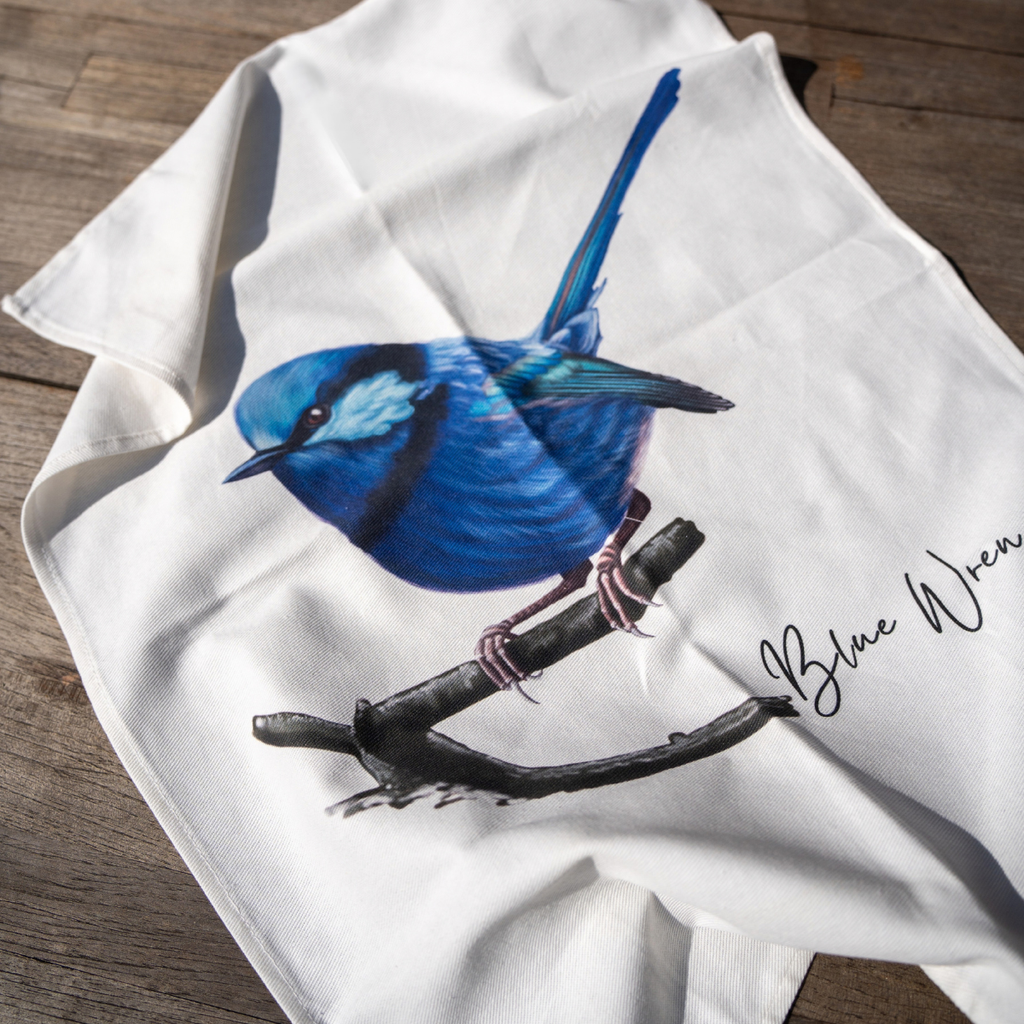 A white kitchen towel featuring a detailed illustration of a blue bird perched on a branch, with the text 'Blue Wren' printed in black script. The towel is laid out on a rustic wooden surface.