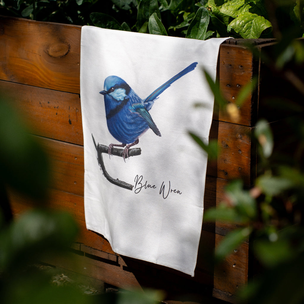 A white kitchen towel draped over a rustic wooden crate, featuring a detailed illustration of a blue bird perched on a branch with the text 'Blue Wren' written below. The scene is framed by soft-focus green foliage in the foreground.