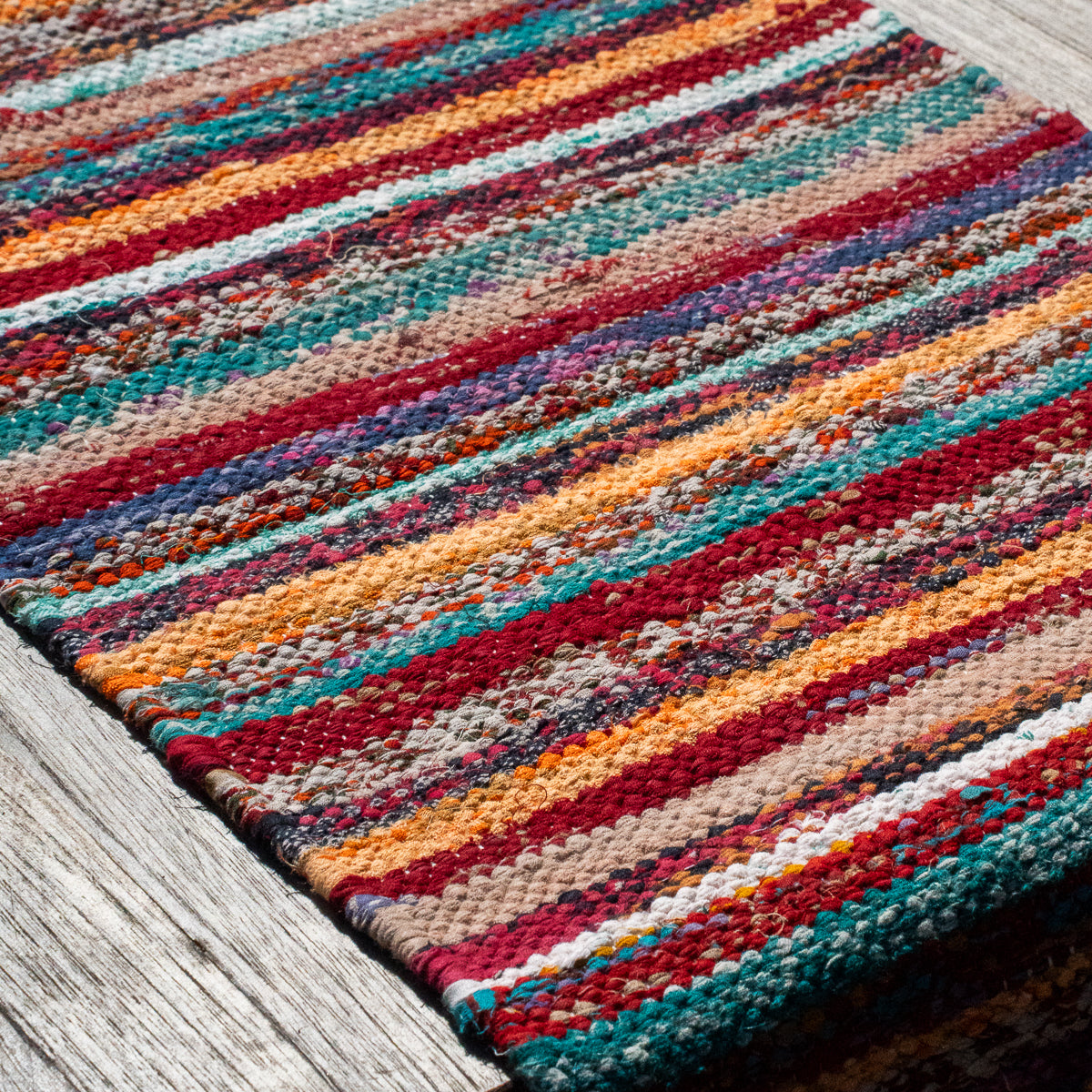 A close-up of a woven area rug with horizontal multicolor stripes in red, orange, teal, beige, and white, laid on a weathered gray wooden surface.