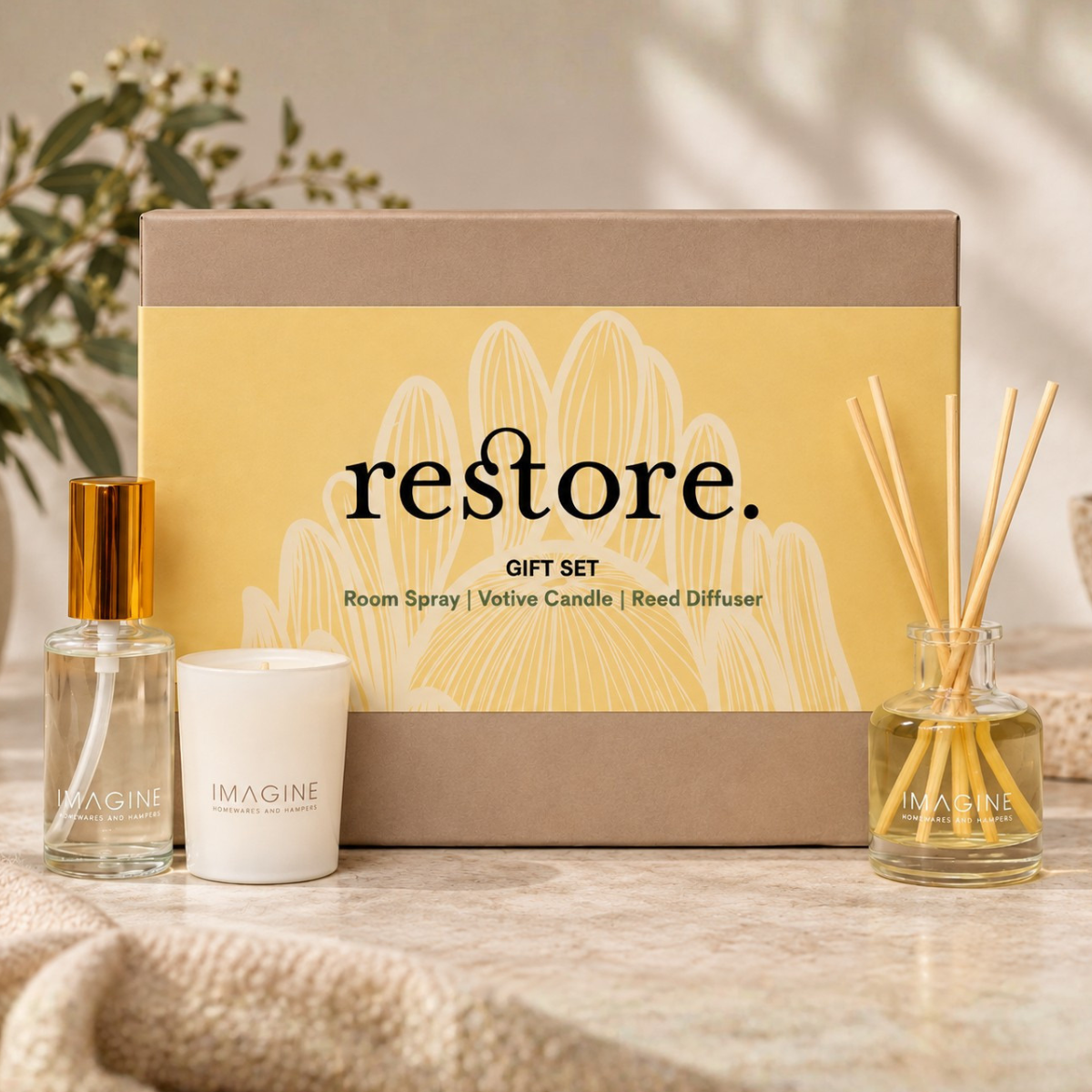 Restore room gift set featuring a yellow and brown box with 'restore.' printed on it, alongside a room spray bottle with a gold cap, a white votive candle, and a glass reed diffuser with wooden sticks.