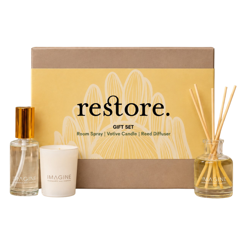 Restore gift set by Imagine Home Fragrance, featuring a room spray bottle with a gold cap, a white votive candle, and a reed diffuser bottle with wooden reeds, arranged in front of a brown and yellow gift box.